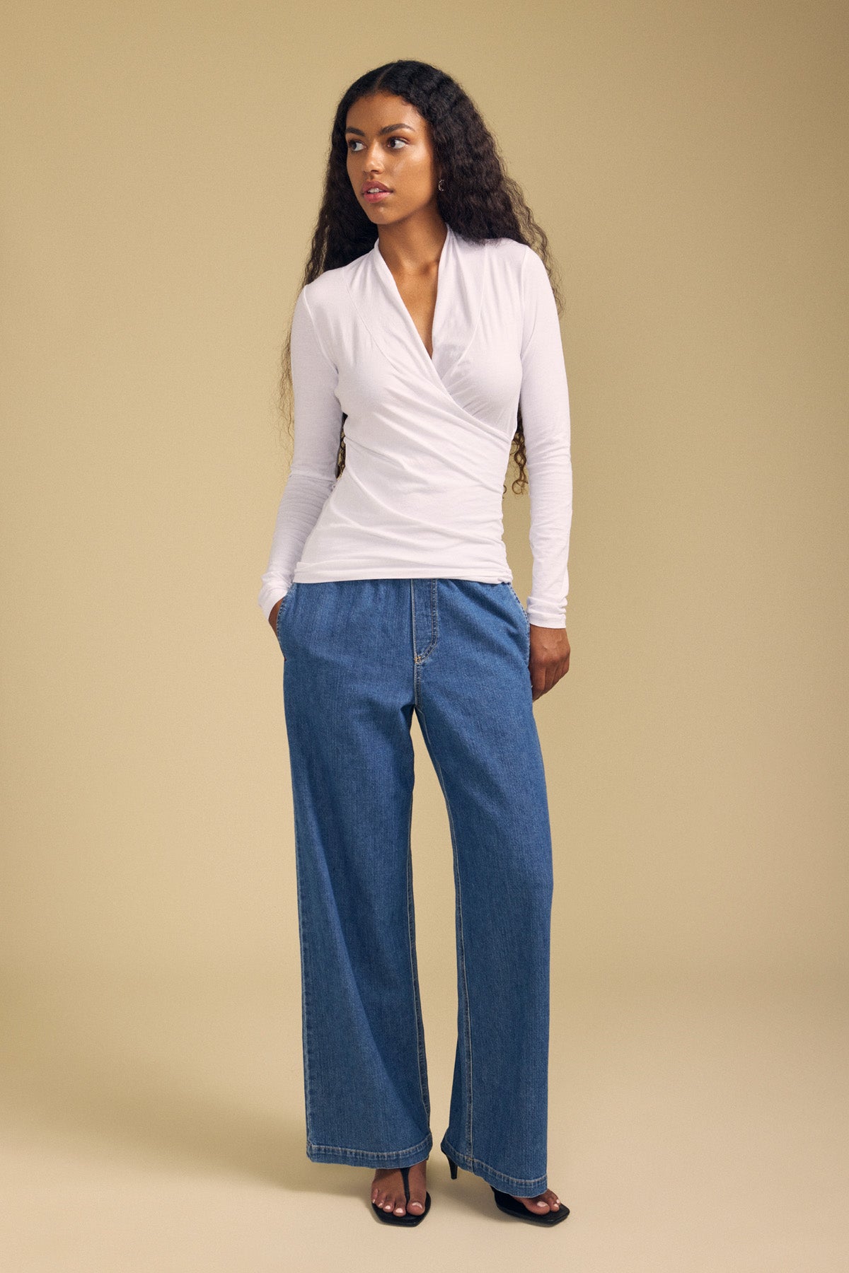 A woman with long curly hair wears the MERI TEE by Velvet by Graham & Spencer—a white cross-over wrap tee with a flattering V-neckline—paired with wide-leg blue jeans and black open-toe heels, standing against a plain beige background.