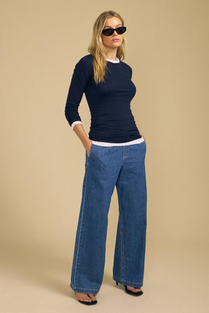 A woman wearing sunglasses and the ZOFINA TEE by Velvet by Graham & Spencer—a fitted long sleeve top with an ultra-soft gauzy whisper feel and classic crew neckline—is styled with white cuffs, wide-leg blue jeans, and black open-toe heels, standing with hands in pockets against a beige background.