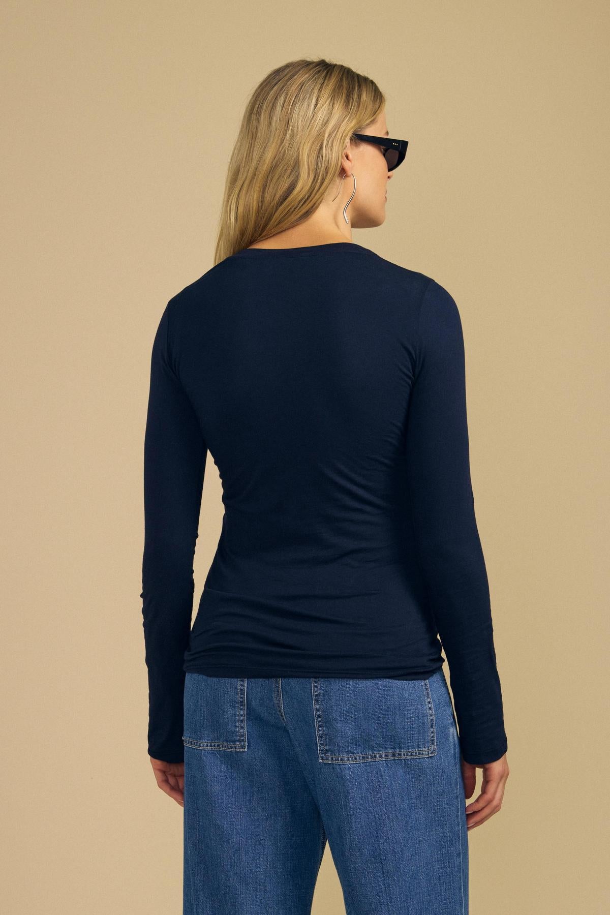 A person with long blonde hair, wearing sunglasses, blue jeans, and the ZOFINA TEE by Velvet by Graham & Spencer—a fitted navy long-sleeve top featuring a classic crew neckline in ultra-soft gauzy whisper fabric—stands facing away from the camera against a beige background.