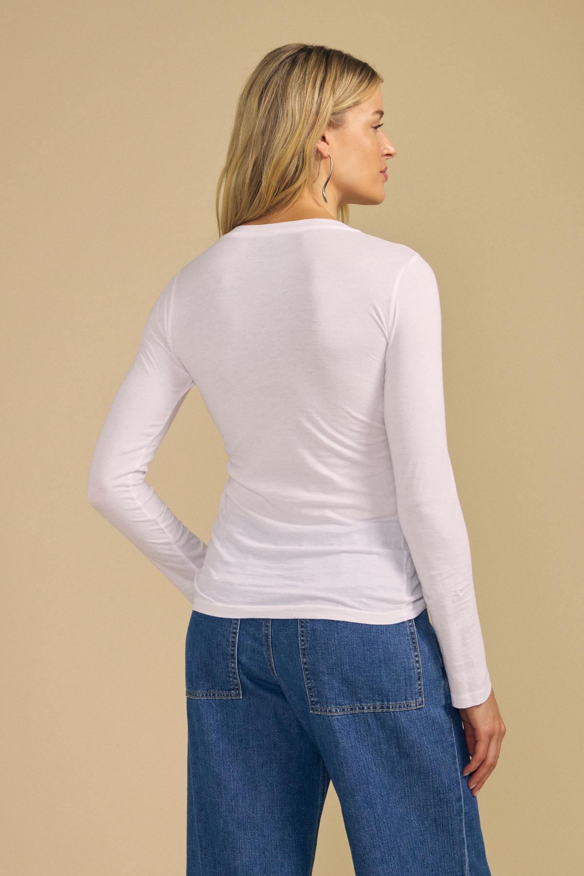A woman stands facing away, wearing the ZOFINA TEE by Velvet by Graham & Spencer—an ultra-soft white fitted long sleeve top with a classic crew neckline—paired with blue jeans against a beige background.-42029125271745