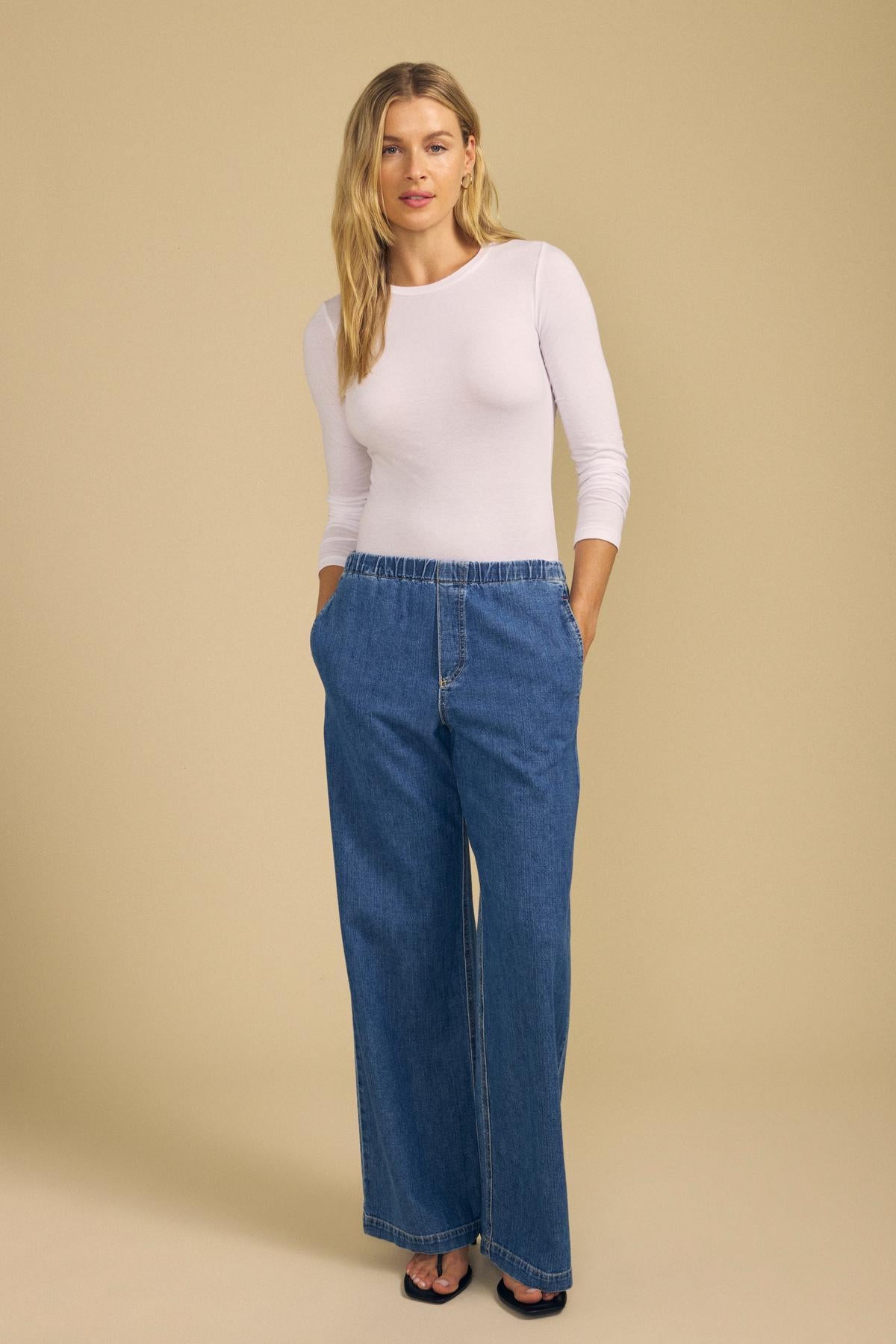 A woman stands against a beige background wearing the ZOFINA TEE by Velvet by Graham & Spencer—an ultra-soft white fitted long sleeve crew neck top—paired with blue wide-leg jeans and black open-toe shoes, with her hands in her pockets.-42029125206209