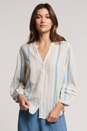 A woman faces forward in the Velvet by Graham & Spencer JUDITH TOP, a lightweight blue and beige striped blouse with a split neckline, paired with a blue skirt against a plain background.