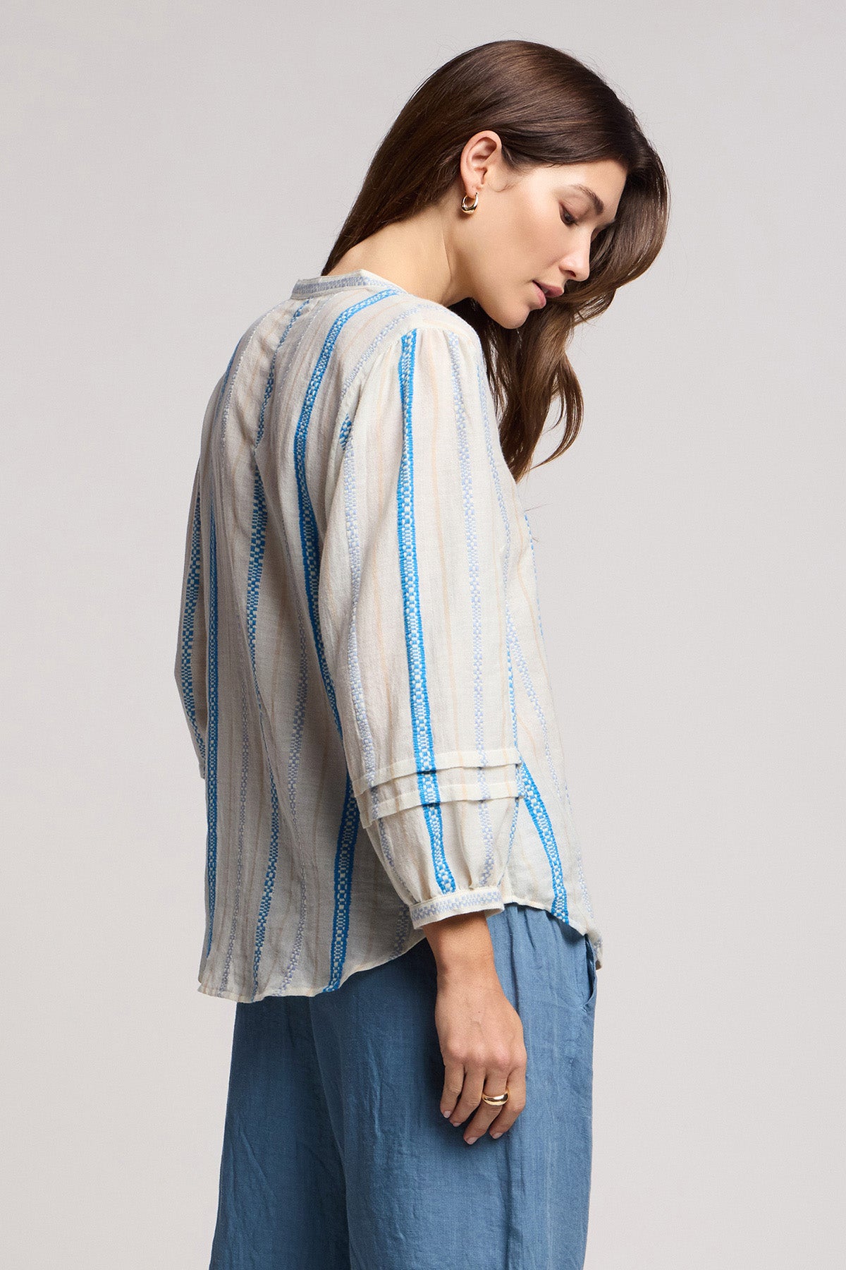 A woman stands sideways against a neutral background, wearing the JUDITH TOP by Velvet by Graham & Spencer—a long-sleeve, light-colored striped cotton jacquard blouse with blue accents—paired with loose blue pants.-41371947565249