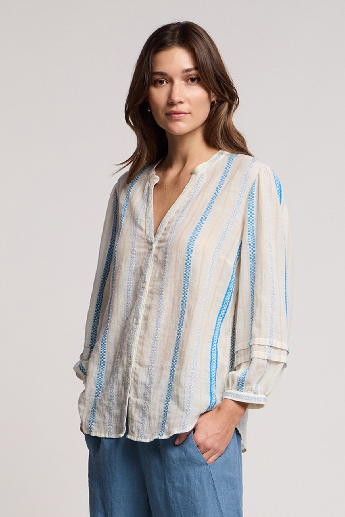 A woman stands facing the camera in the JUDITH TOP by Velvet by Graham & Spencer, a lightweight blouse with a split neckline and blue and beige accents, paired with light blue pants against a plain background.-41371948581057