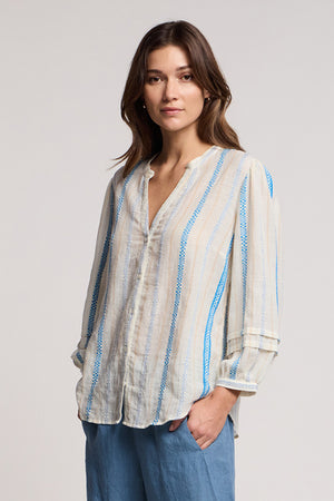 A woman stands facing the camera in the JUDITH TOP by Velvet by Graham & Spencer, a lightweight blouse with a split neckline and blue and beige accents, paired with light blue pants against a plain background.