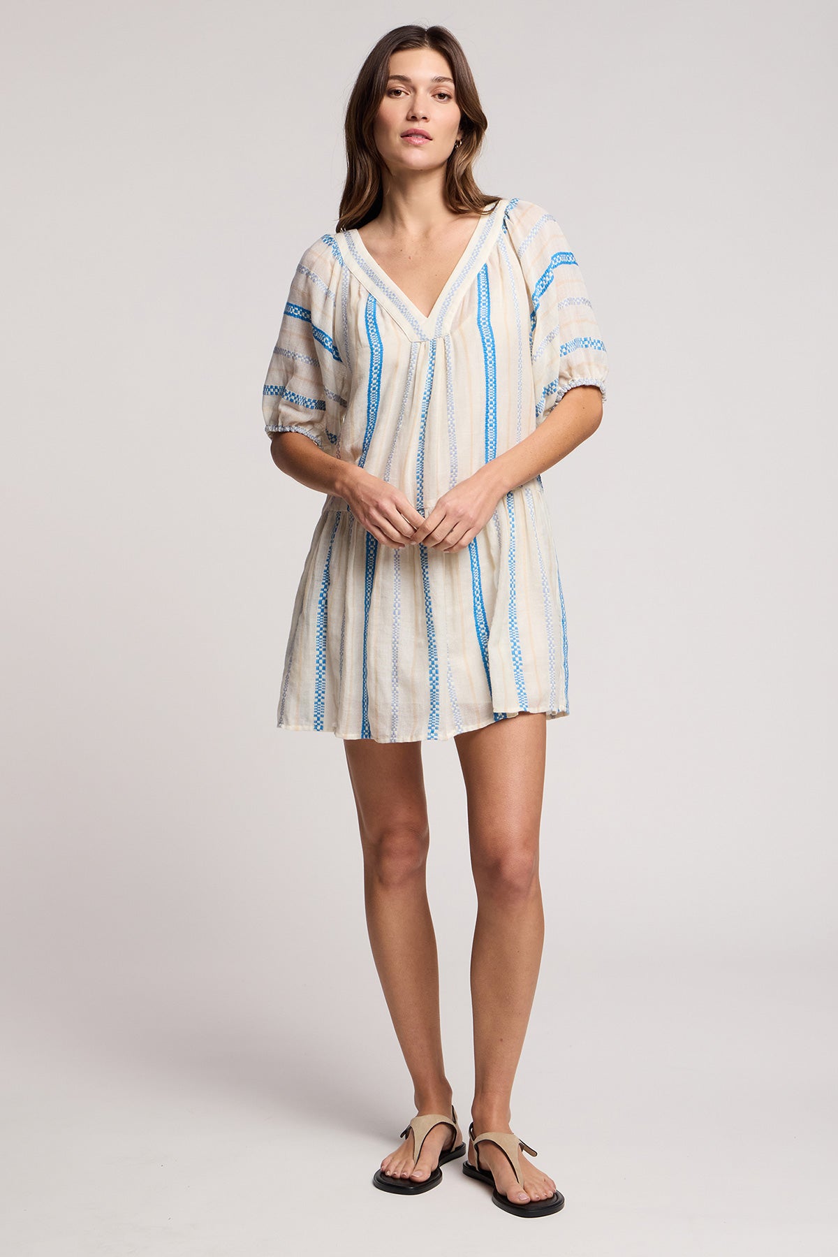 A woman in the SELITA DRESS by Velvet by Graham & Spencer— a short, loose-fitting cotton jacquard mini dress with blue vertical stripes and puff sleeves—stands against a plain background, paired with black sandals.