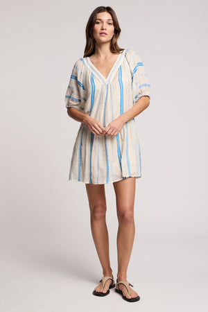 A woman in the SELITA DRESS by Velvet by Graham & Spencer— a short, loose-fitting cotton jacquard mini dress with blue vertical stripes and puff sleeves—stands against a plain background, paired with black sandals.