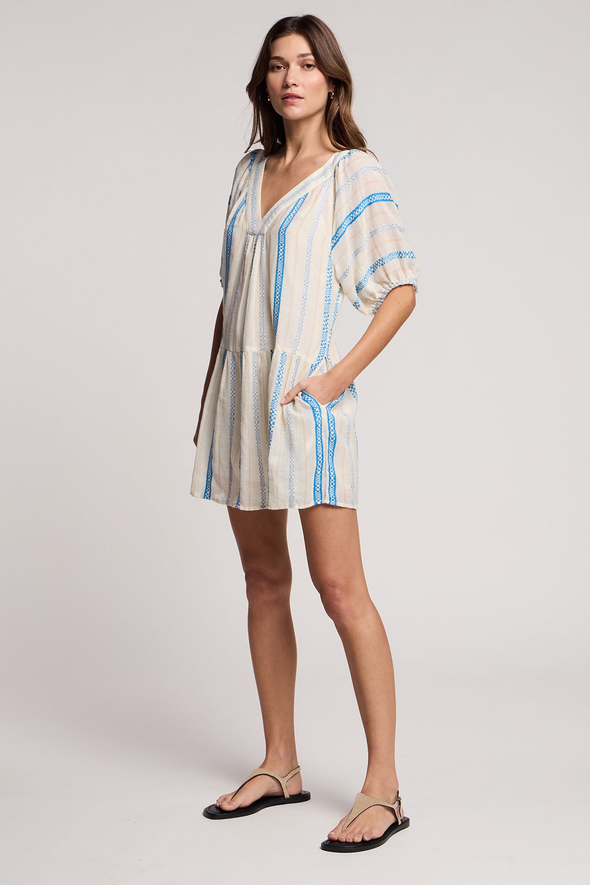 A woman models the SELITA DRESS by Velvet by Graham & Spencer— a cream jacquard cotton romper with blue stripes and pockets—paired with black sandals, set against a plain light background.