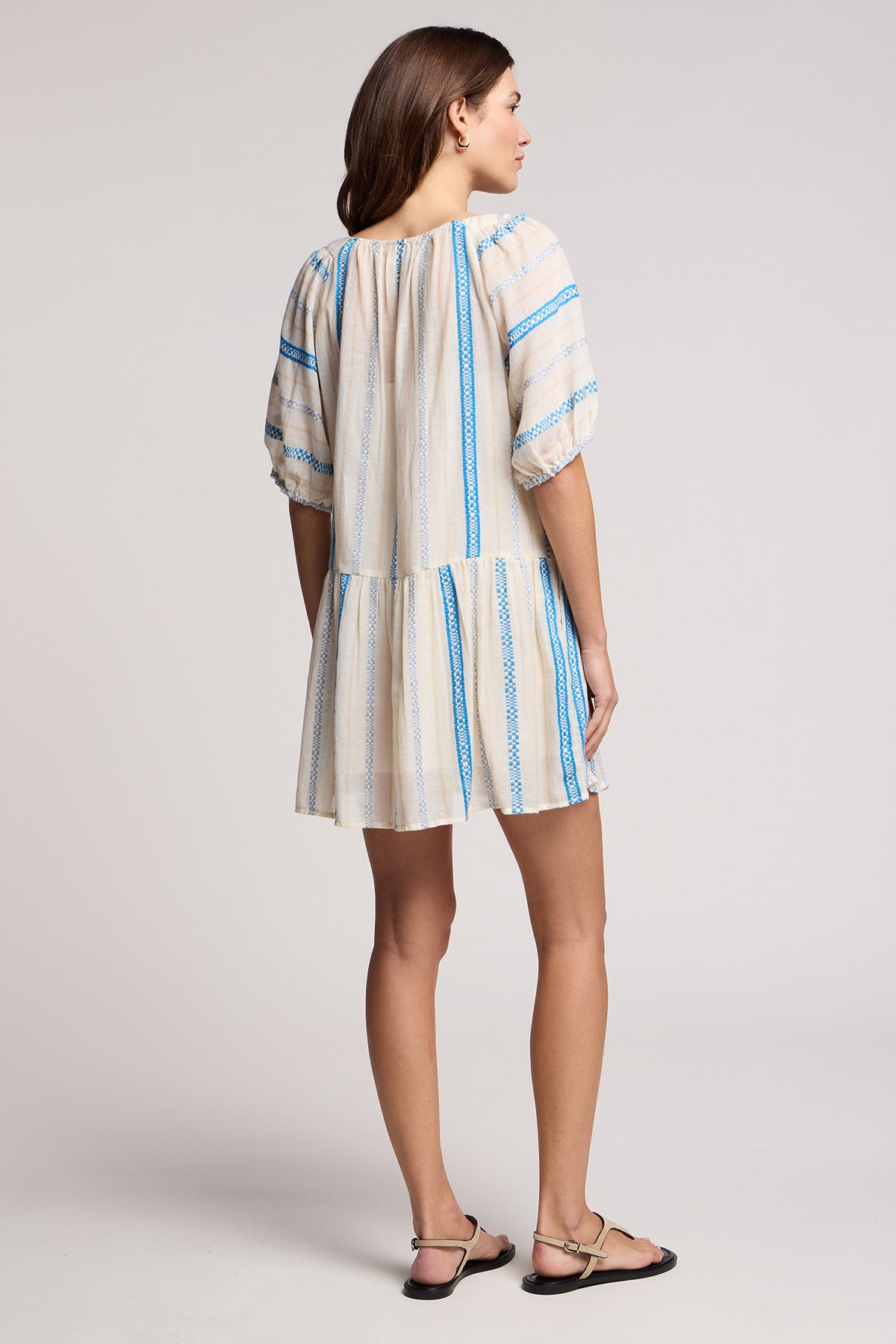 A woman faces away in the SELITA DRESS by Velvet by Graham & Spencer—a loose, light-hued cotton jacquard mini dress with blue stripes and short puff sleeves—paired with black sandals.-41374563991745