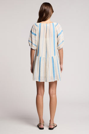 A woman stands with her back to the camera in the SELITA DRESS by Velvet by Graham & Spencer, a loose cotton jacquard mini dress with blue stripes and puff sleeves, paired with flat sandals against a plain background.
