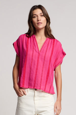 A person wearing the Velvet by Graham & Spencer TALLULAH TOP, a bright pink split neck top with vertical textured stripes and a boxy silhouette, paired with white pants, stands against a plain light background.