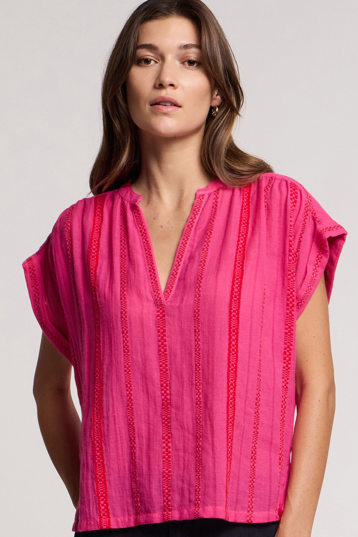A woman wears the Velvet by Graham & Spencer TALLULAH TOP, a bright pink geometric cotton jacquard split neck top with red vertical stitched stripes, standing against a neutral background.-41372222521537