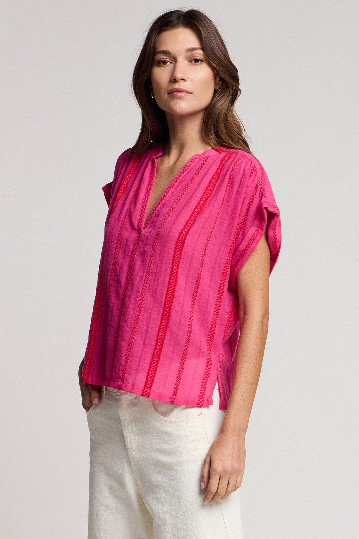 A woman wears the TALLULAH TOP by Velvet by Graham & Spencer—a bright pink, split neck, vertically striped top with a boxy silhouette—paired with off-white pants against a plain background.