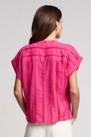 A woman with long brown hair is seen from the back wearing the TALLULAH TOP by Velvet by Graham & Spencer—a bright pink cap sleeve style with red stitched stripes—paired with off-white bottoms.
