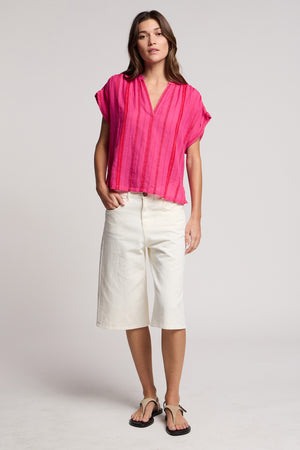 A woman stands facing forward against a plain light background, wearing the TALLULAH TOP in bright pink by Velvet by Graham & Spencer, paired with white knee-length shorts and black sandals.