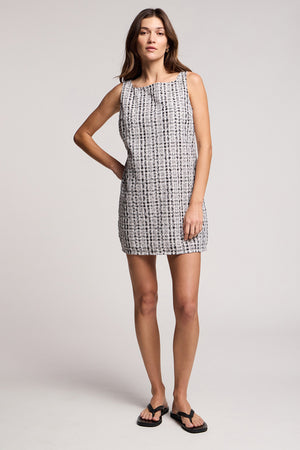A woman stands in a studio wearing the KATSY DRESS by Velvet by Graham & Spencer— a sleeveless, black-and-white gingham mini dress—paired with black sandals against a plain background.