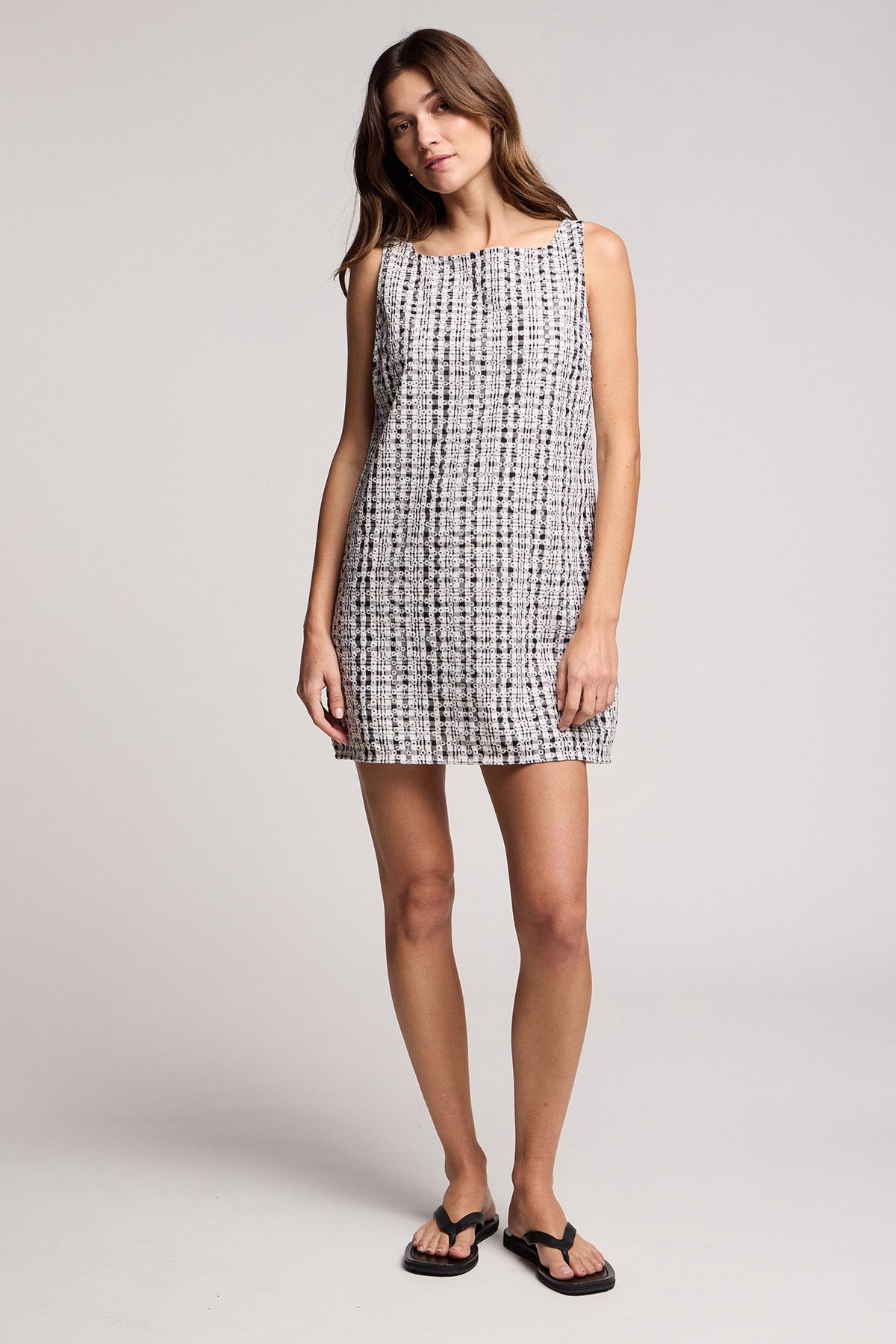 A woman poses in a studio wearing the KATSY DRESS by Velvet by Graham & Spencer—a sleeveless black and white gingham mini dress—paired with black sandals.