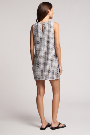 A woman faces away, wearing the sleeveless KATSY DRESS by Velvet by Graham & Spencer—a patterned gingham mini dress—paired with black sandals against a plain background.