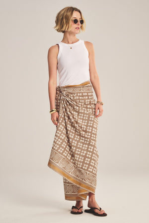 A woman wearing sunglasses, a white sleeveless top, the Velvet by Graham & Spencer GRECHEN SARONG skirt, black sandals, and gold jewelry stands against a plain background.