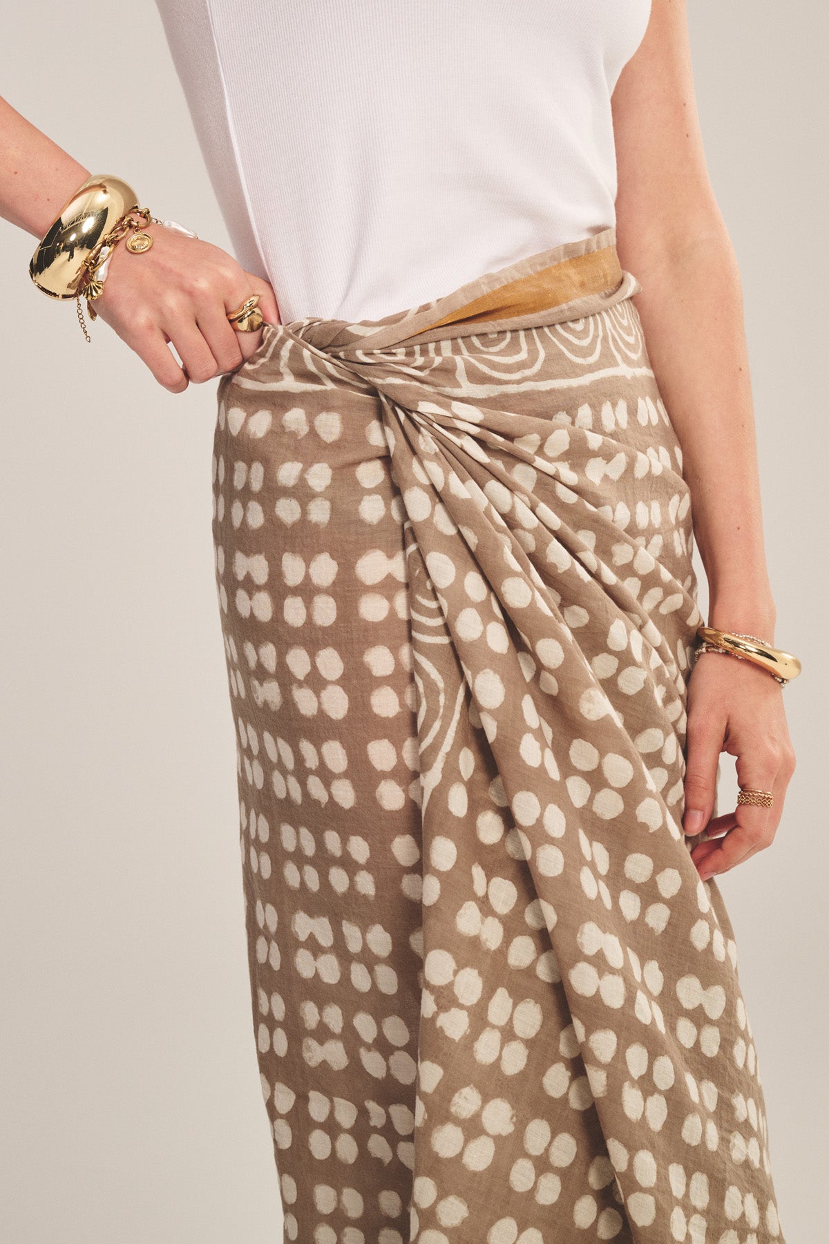 Model wears the GRECHEN SARONG by Velvet by Graham & Spencer—a beige printed wrap—paired with a white sleeveless top and gold bracelets and rings, set against a plain backdrop.