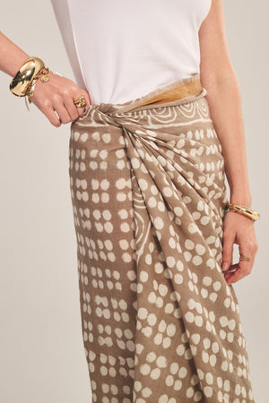 Model wears the GRECHEN SARONG by Velvet by Graham & Spencer—a beige printed wrap—paired with a white sleeveless top and gold bracelets and rings, set against a plain backdrop.