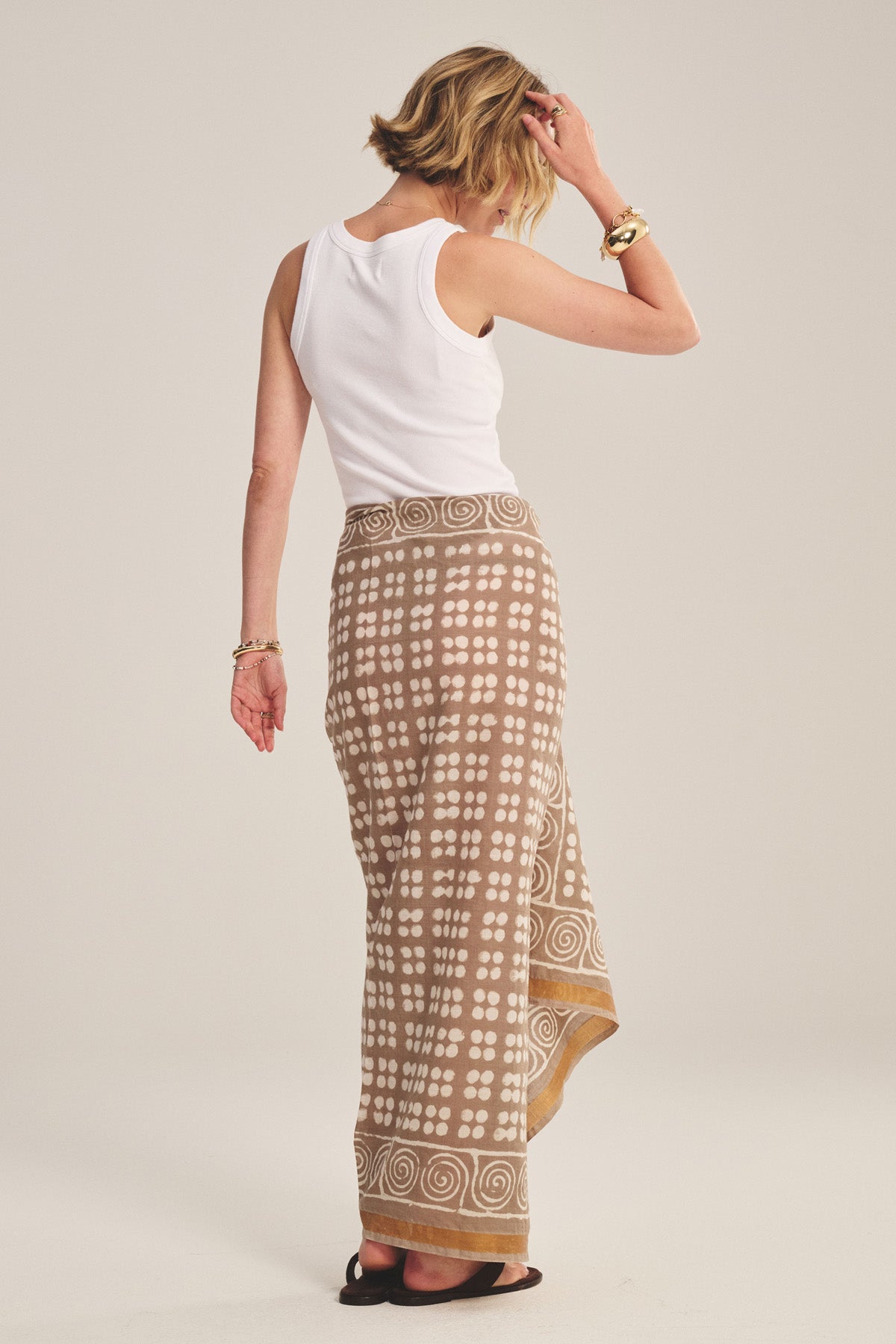 A woman, facing away, wears a white sleeveless top, black sandals, gold bracelets, and the GRECHEN SARONG by Velvet by Graham & Spencer—a beige printed wrap skirt—with her left hand touching her hair.