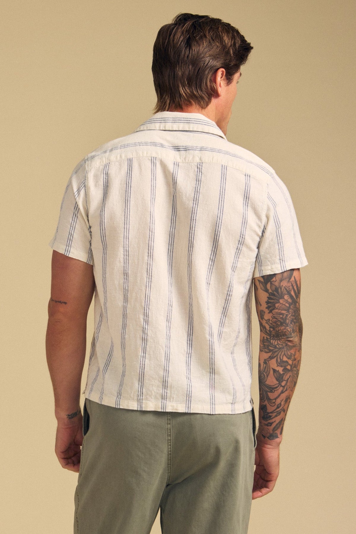 A man stands with his back to the camera, wearing the GRIFFEN SHIRT by Velvet by Graham & Spencer—a striped, short-sleeve, camp-collar shirt in a cotton-linen blend—paired with light green pants.-41626558562497
