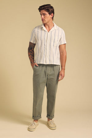 A man stands against a beige background wearing Velvet by Graham & Spencer’s BRANSON PANT—straight-leg, olive green twill with an elastic drawstring waist—paired with a short-sleeve striped shirt and beige sneakers, looking to the side.