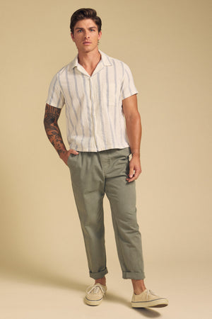 A man with a left forearm tattoo stands against a beige background wearing Velvet by Graham & Spencer’s GRIFFEN SHIRT—a striped, short-sleeve, camp collar button-up in cotton-linen—paired with olive pants and cream sneakers.