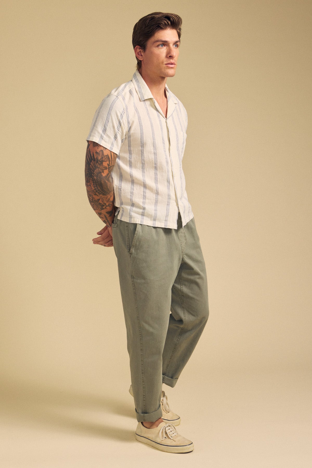 A man stands with hands behind his back, wearing a short-sleeve striped shirt, Velvet by Graham & Spencer's BRANSON PANT in light green cotton sanded twill, and beige shoes, against a plain beige background.