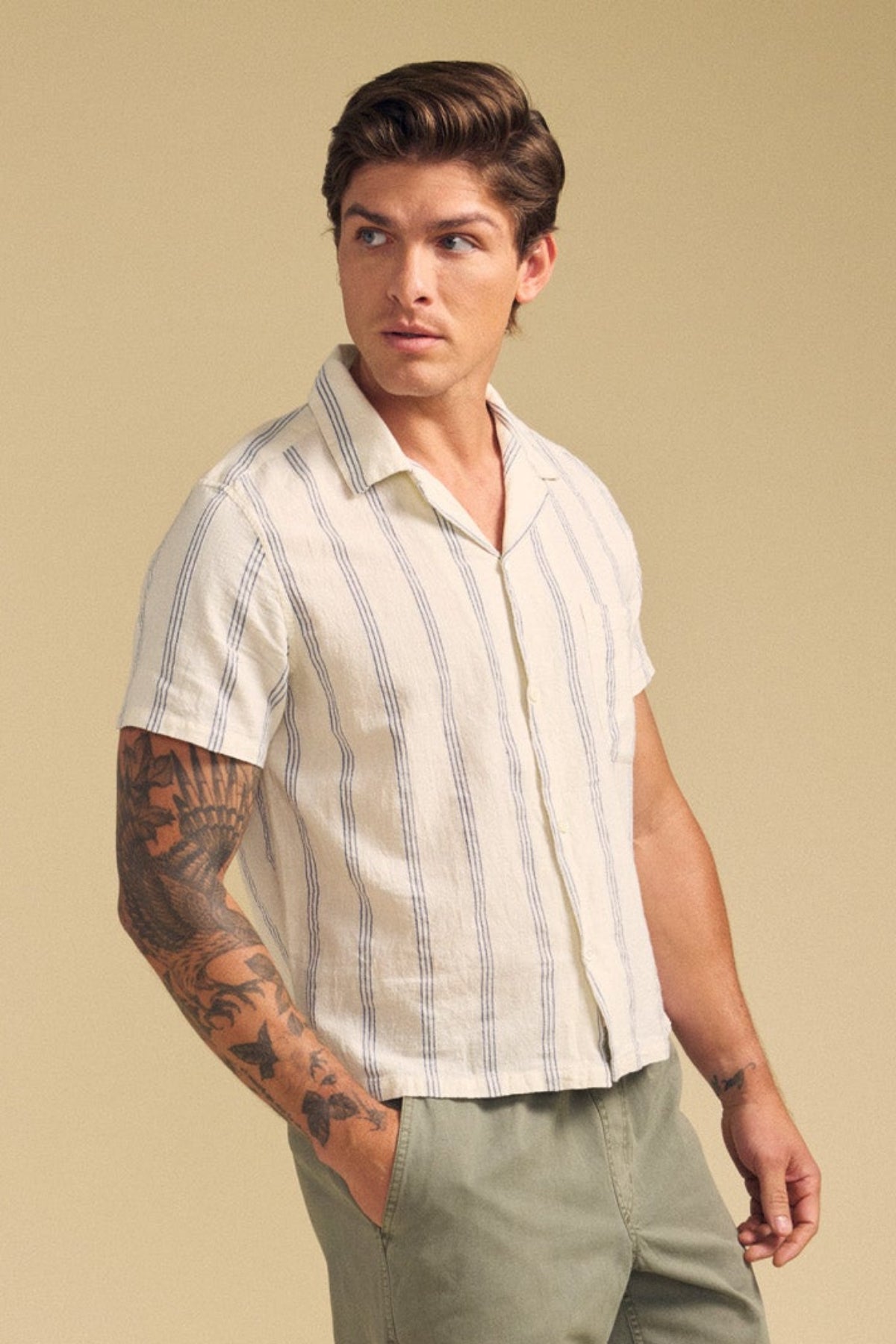 A man models the GRIFFEN SHIRT by Velvet by Graham & Spencer—a striped, camp-collar, short-sleeve cotton-linen blend—paired with olive pants. He stands against a beige background, showing a tattoo on his left forearm and glancing sideways.