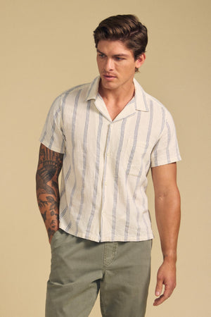 A man wears the GRIFFEN SHIRT by Velvet by Graham & Spencer—a cotton-linen blend, striped, short-sleeve camp collar shirt—paired with olive-green pants, standing against a plain beige background.