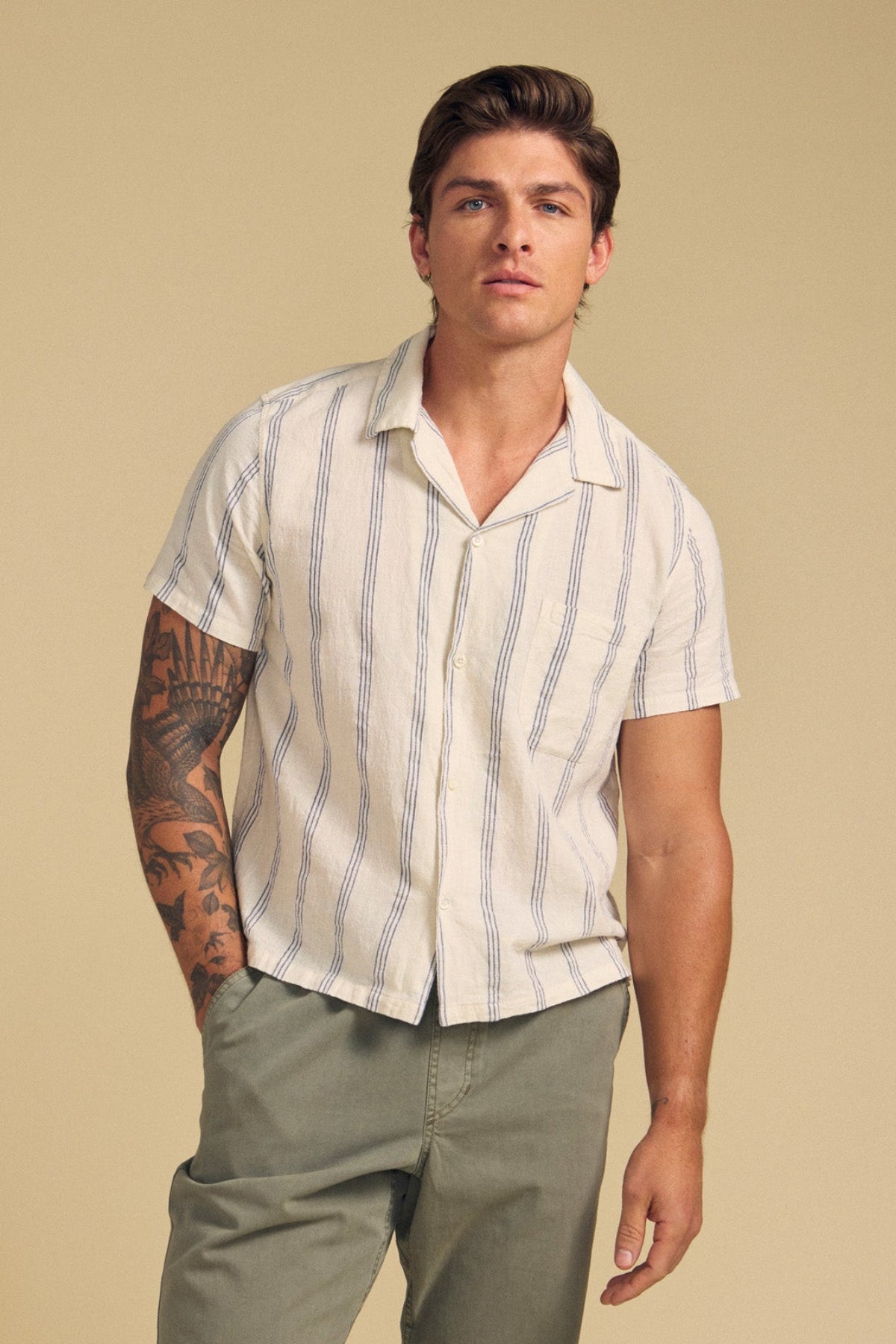 A man with a tattooed arm wears the Velvet by Graham & Spencer GRIFFEN SHIRT—a cream-striped, camp-collar short-sleeve in soft cotton-linen blend—paired with olive-green pants, standing against a beige background.-41626558955713