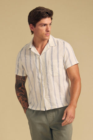 A man wears the GRIFFEN SHIRT by Velvet by Graham & Spencer, a striped camp-collar short-sleeve style in a cotton-linen blend, paired with light olive pants and standing against a beige background.