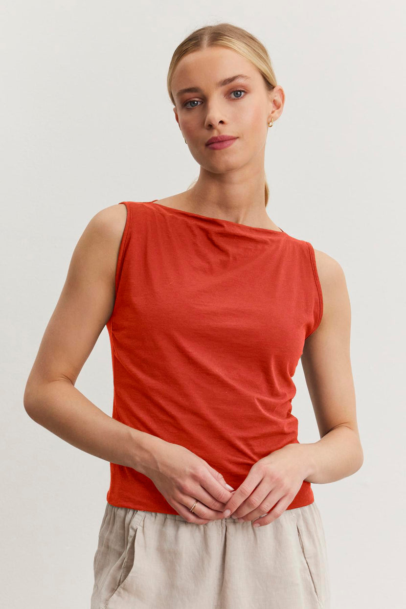 A woman with blonde hair tied back, wearing a sleeveless red top and light beige pants, stands against a plain white background. She looks directly at the camera with a neutral expression, showcasing the EMILIA TANK TOP from Velvet by Graham & Spencer in modern minimal design made from soft cotton.