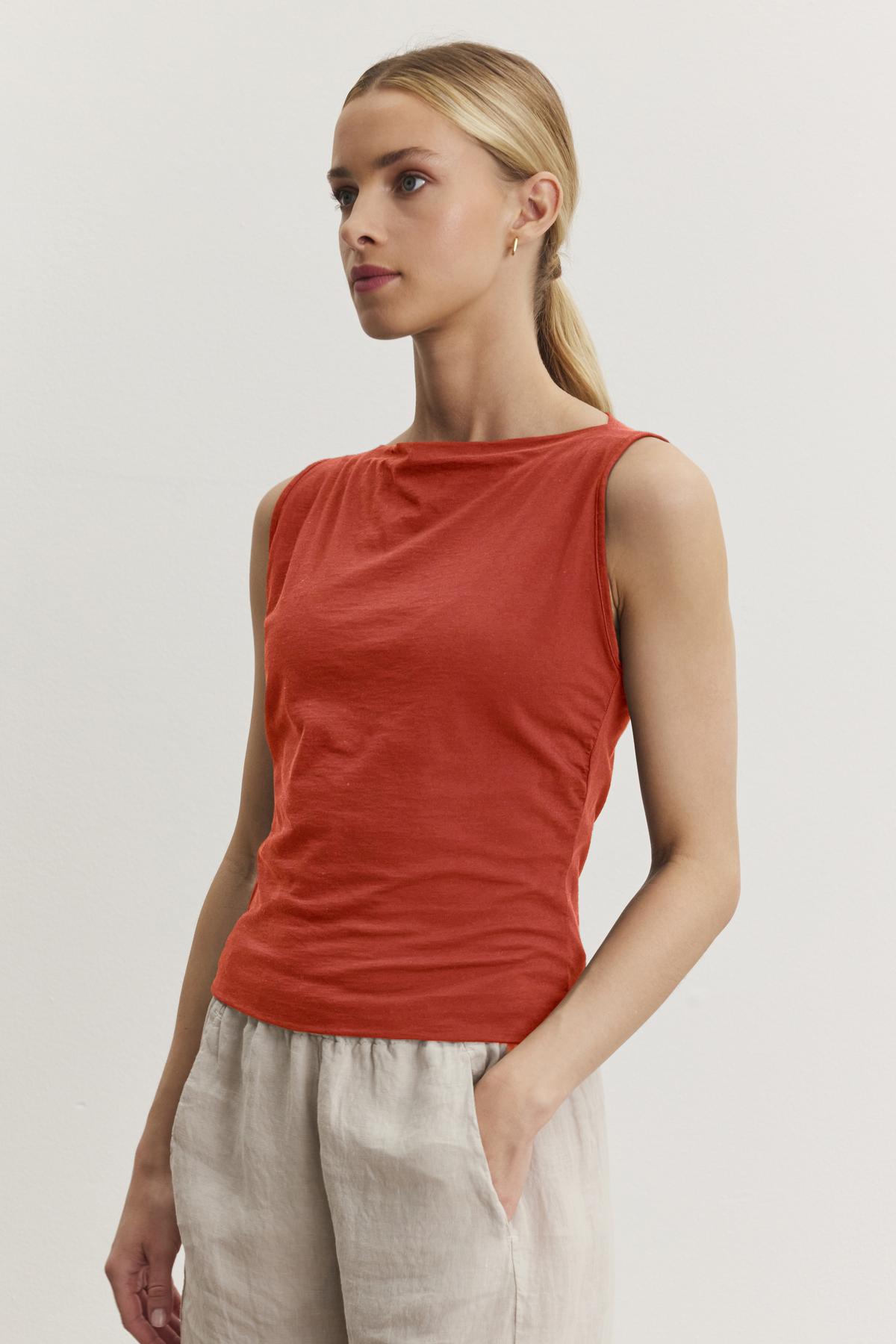   A person with blonde hair tied back is wearing a sleeveless, red EMILIA TANK TOP made of soft cotton and beige pants, standing against a plain, white background—perfect for casual wear with a modern minimal design by Velvet by Graham & Spencer. 