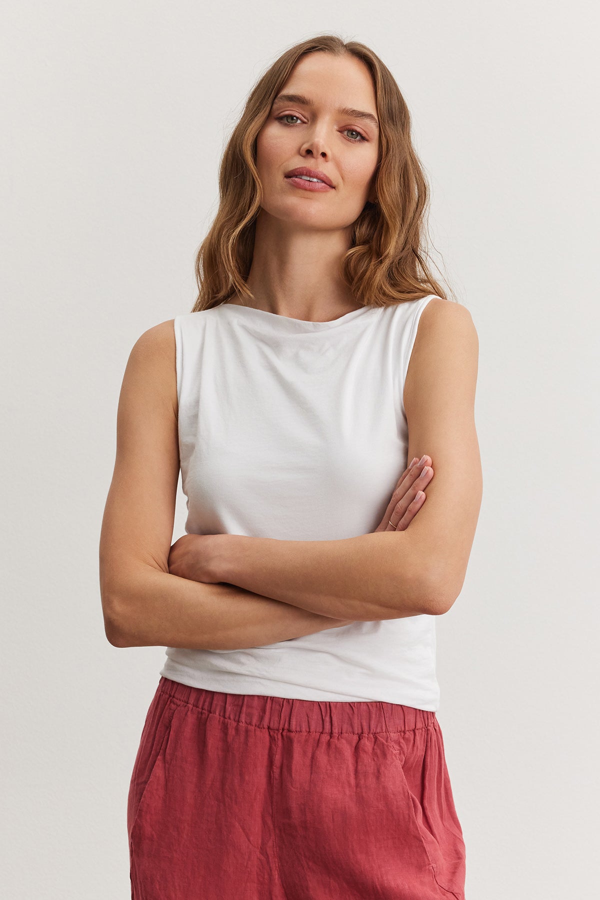   A woman with medium-length hair, wearing a Velvet by Graham & Spencer EMILIA TANK TOP and red pants, stands with her arms crossed. 