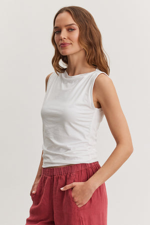 A woman in a white Emilia tank top and red pants stands with one hand in her pocket, looking slightly to the side against a plain background.