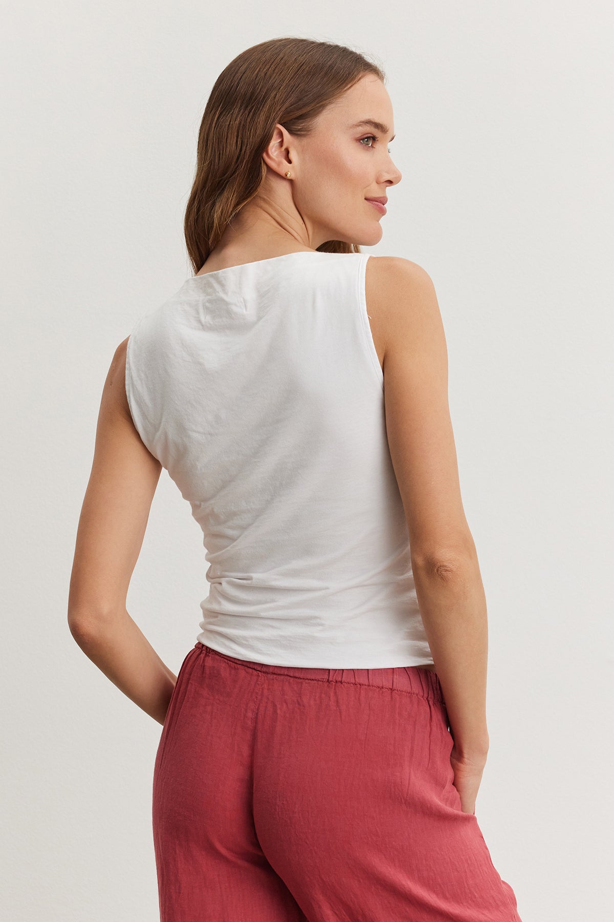   A woman in a Velvet by Graham & Spencer EMILIA TANK TOP and red pants, viewed from behind, looking over her shoulder with a neutral expression. 