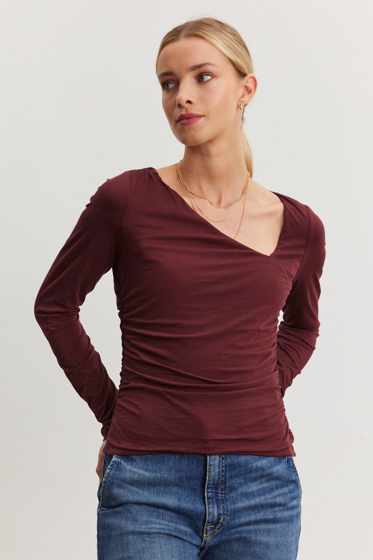 A person wearing the FIONA TEE by Velvet by Graham & Spencer, a maroon long-sleeve top with an asymmetrical neckline, paired with blue jeans, stands against a plain background.-38483582451905