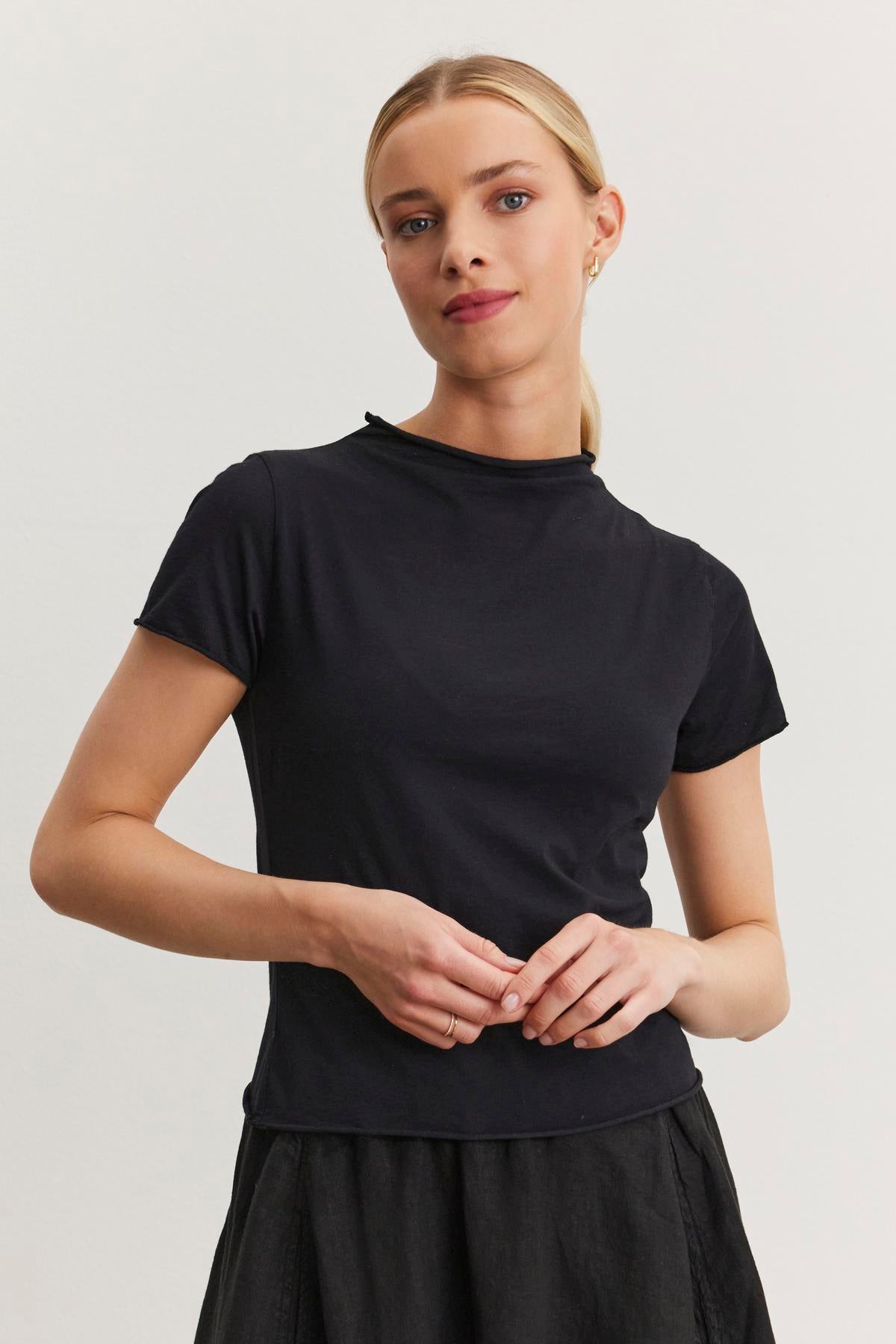 A person with blonde hair stands against a plain background, wearing the Velvet by Graham & Spencer JACKIE MOCK NECK TEE in gauzy whisper fabrication and a black skirt, with hands loosely clasped.-37104257761473