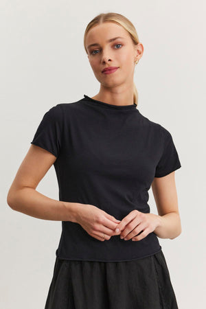 A person with blonde hair stands against a plain background, wearing the Velvet by Graham & Spencer JACKIE MOCK NECK TEE in gauzy whisper fabrication and a black skirt, with hands loosely clasped.