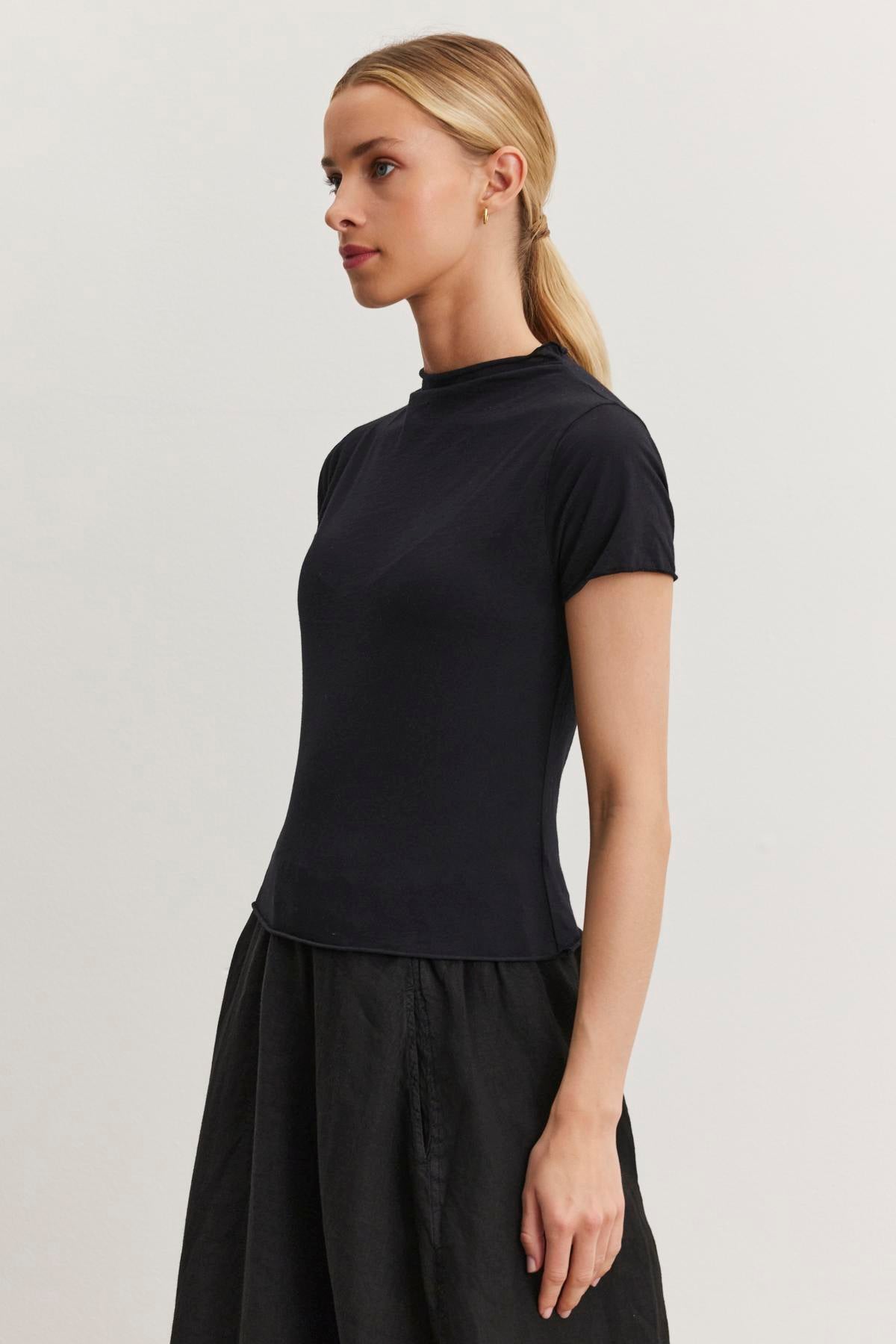   A woman with blonde hair tied back in a ponytail is wearing a black JACKIE MOCK NECK TEE by Velvet by Graham & Spencer and a matching black skirt, both showcasing a fitted cropped silhouette, standing against a plain white background. 