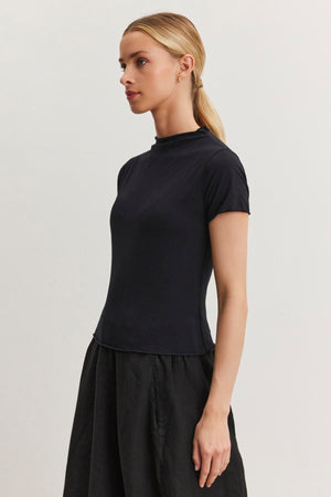 A woman with blonde hair tied back in a ponytail is wearing a black JACKIE MOCK NECK TEE by Velvet by Graham & Spencer and a matching black skirt, both showcasing a fitted cropped silhouette, standing against a plain white background.