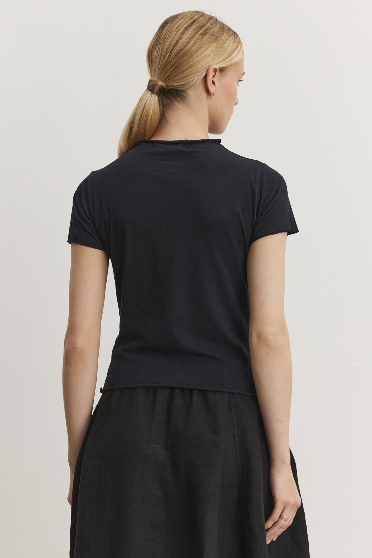   A person with blonde hair tied in a low ponytail is standing with their back to the camera, wearing a fitted cropped black JACKIE MOCK NECK TEE by Velvet by Graham & Spencer and a black skirt. 