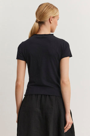 A person with blonde hair tied in a low ponytail is standing with their back to the camera, wearing a fitted cropped black JACKIE MOCK NECK TEE by Velvet by Graham & Spencer and a black skirt.