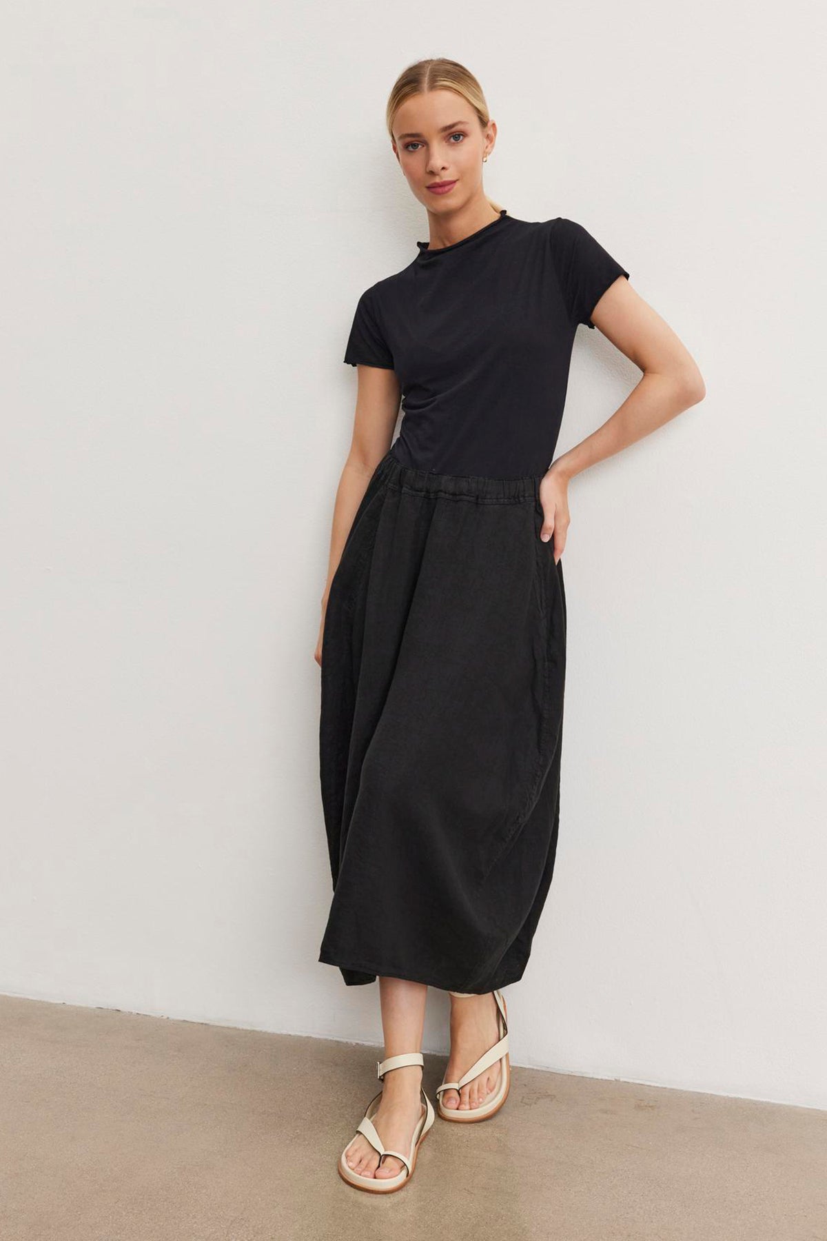 A woman in a black JACKIE MOCK NECK TEE by Velvet by Graham & Spencer and black midi skirt stands against a white wall, white sandals adorning her feet. She is leaning to her left with her hand casually slipped in her pocket.-37104257794241