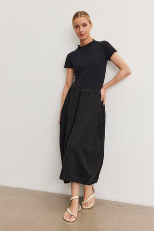 A woman in a black JACKIE MOCK NECK TEE by Velvet by Graham & Spencer and black midi skirt stands against a white wall, white sandals adorning her feet. She is leaning to her left with her hand casually slipped in her pocket.