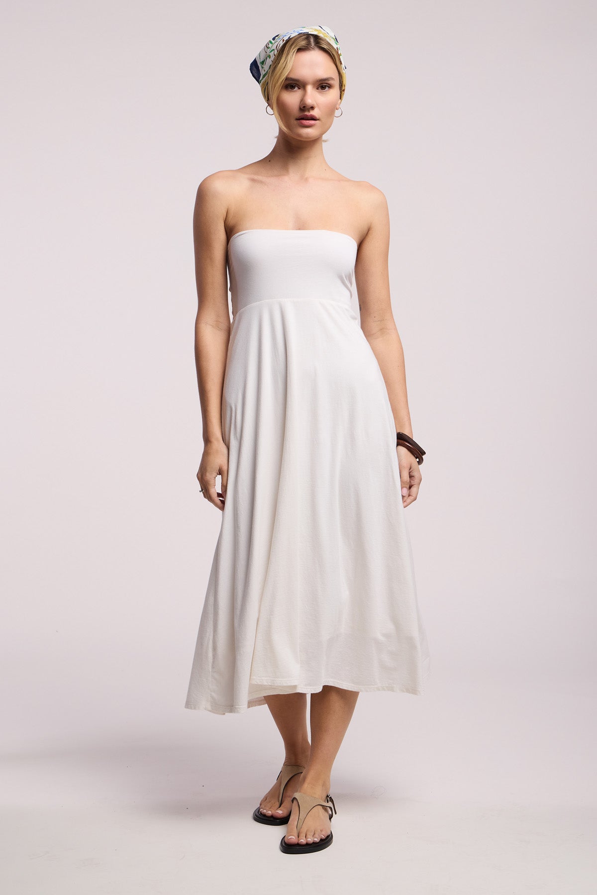 A woman faces forward in a Velvet by Graham & Spencer LUELLA SKIRT, paired with sandals, a headscarf, hoop earrings, and bracelet against a plain light backdrop.