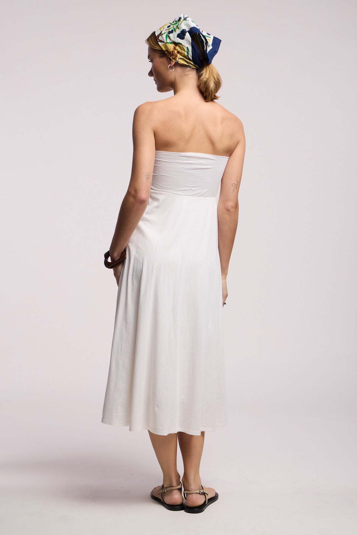 A woman wearing the LUELLA SKIRT by Velvet by Graham & Spencer, styled with black sandals and a patterned headscarf, stands facing away from the camera against a plain background.-41371962278081